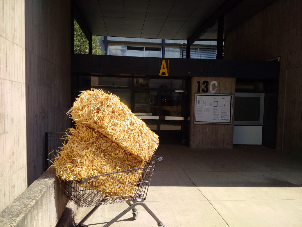 two haybails in a shopping cart in front of a brutalist building